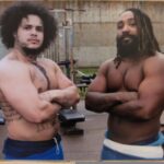 malik and another prisoner stand shirtless looking strong with arms crossed.