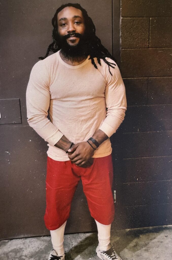 Malik smiles at the camera while standing with hands folded, wearing a white shirt and red shorts.