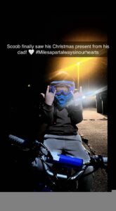 Amir wears a full protection helmet and sits on a small dirt bike while excitedly throwing horns with both hands, text reads "Scoob finally saw his Christmas present from his dad! heart emoji, #MilesApartAlwaysInOurHearts