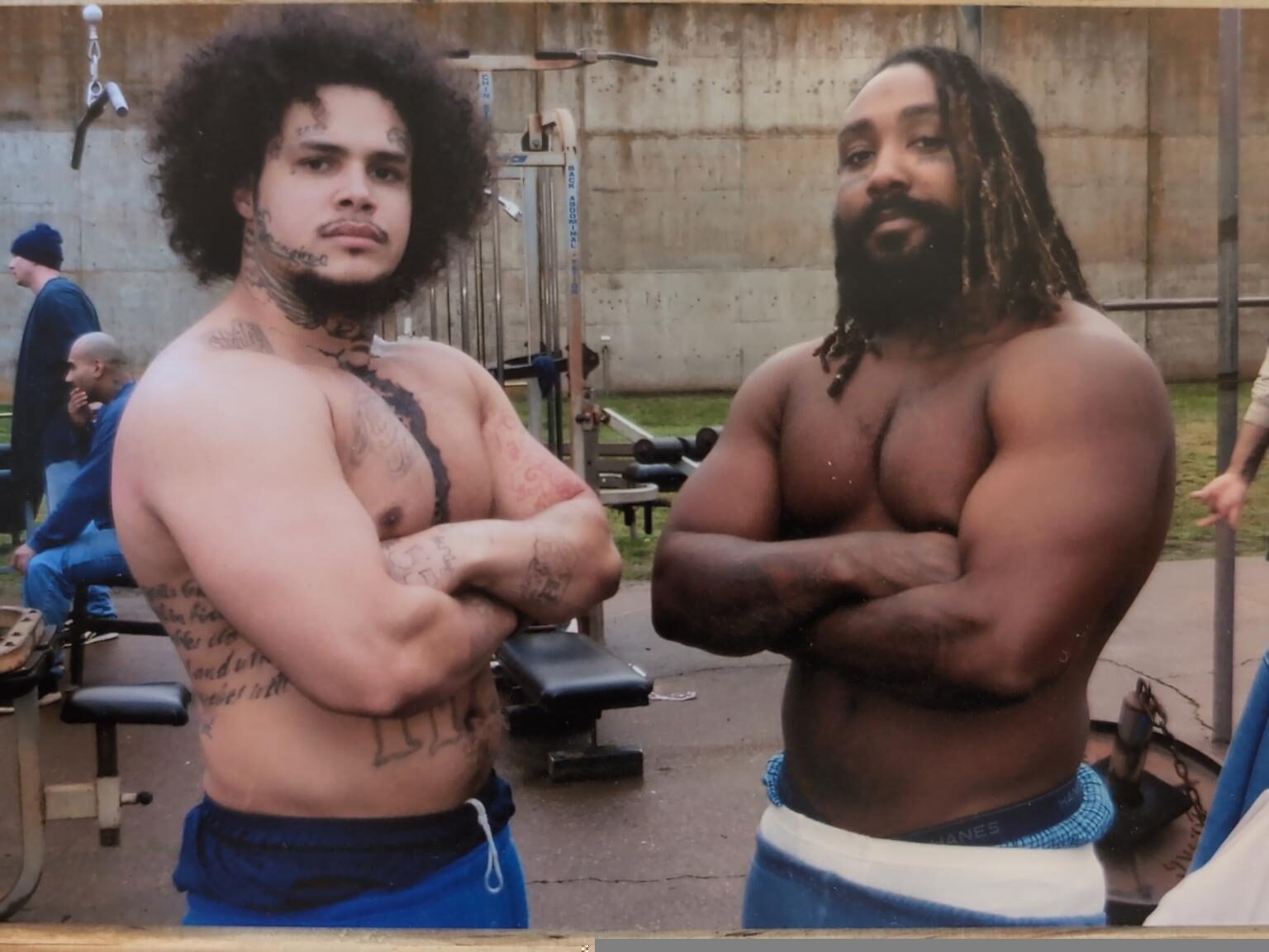 malik and another prisoner stand shirtless looking strong with arms crossed.